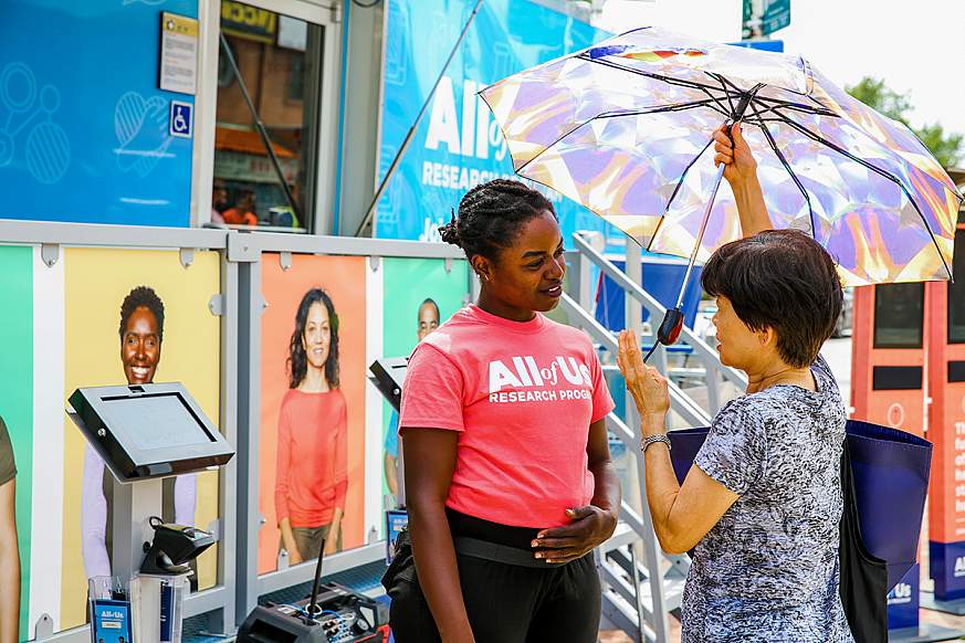 Young woman with All of Us tee shirt speaks to woman holding an umbrella.