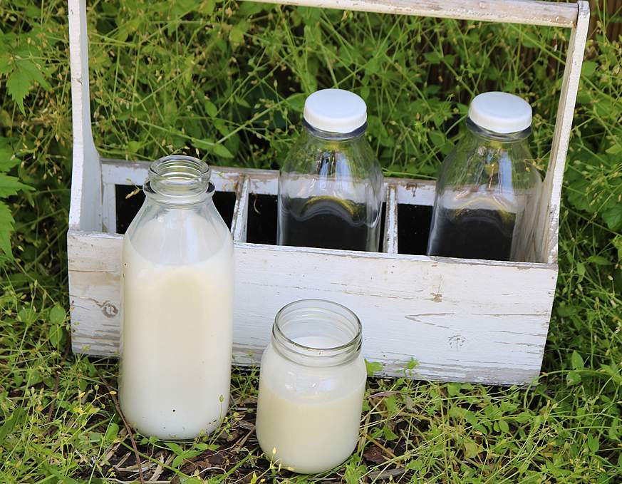 The image shows two bottles of raw milk in glass bottles in a wooden bottle holder placed on the ground.