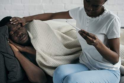 A young woman reading a thermometer and checking the forehead of a sick young man on a couch.