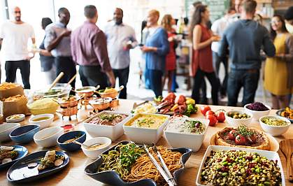 A table displaying a variety of foods with a crowd of people standing around and talking behind it.