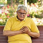 A senior man sitting on a park bench and holding his chest in pain