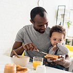 Father with young son on his lap spreading peanut butter on toast.