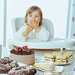 Young toddler in a high chair eating chocolate cake, with an assortment of pastries and cakes on the table in the foreground.