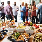 A table displaying a variety of foods with a crowd of people standing around and talking behind it.