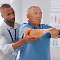 An older man in physical therapy lifting small weights with help from a therapist.