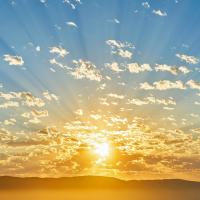 A sunrise over a mountain range with rays of sunlight streaming through scattered clouds in a blue sky.